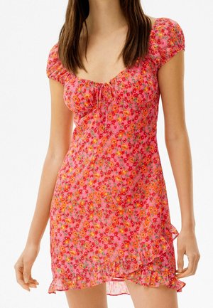 Mujer con un vestido corto de manga corta, estampado floral rosa, con dobladillo con volantes y detalle de lazo en el escote, sobre un fondo blanco.