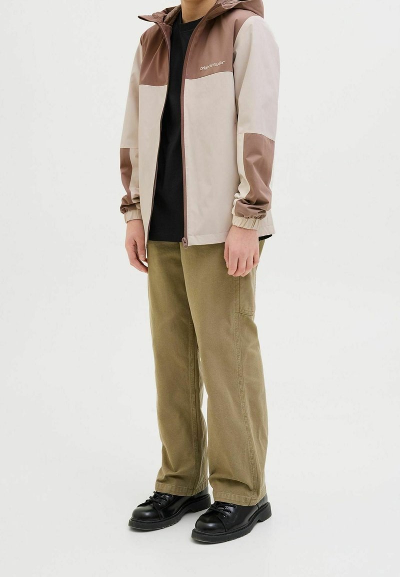 Giacca chiara con dettagli marroni, polsini elastici e cappuccio. Abbinata a pantaloni khaki e scarpe nere. Design e texture semplici.