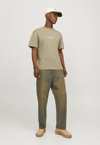 Khaki Baumwoll-T-Shirt mit kurzen Ärmeln und kleinem weißen Text auf der Brust, kombiniert mit lockeren Denim-Jeans mit einer verblassten, mehrfarbigen Textur.