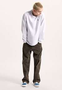 Grijs T-shirt met lange mouwen, losvallende donkerbruine broek, blauwe en witte sneakers, met een klein logo op het shirt.