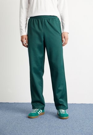 Pantalon de survêtement vert avec un waistband élastique, des poches latérales et une texture lisse, associé à des chaussures de sport vertes avec des rayures blanches.