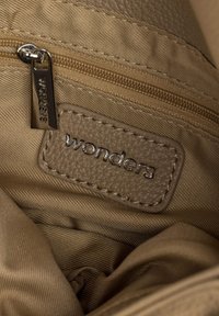 Zbliżenie wnętrza beżowej torebki, pokazujące skórzaną łatkę i suwak z wygrawerowaną nazwą marki "wonders."