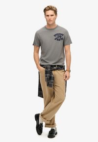 Camiseta de algodón gris con logo azul marino, chinos beige y una camisa de cuadros atada a la cintura. Zapatos negros con calcetines blancos completan el conjunto.