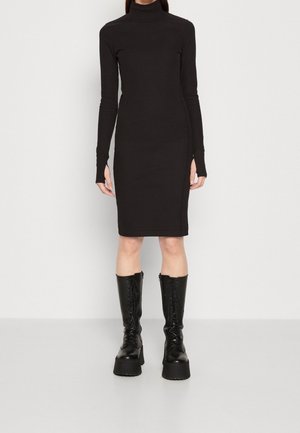 Kobieta ubrana w dopasowaną, długorękawową czarną sukienkę z golfem oraz czarne, platformowe, sięgające do kolan sznurowane botki, stojąca na jednolitym tle.