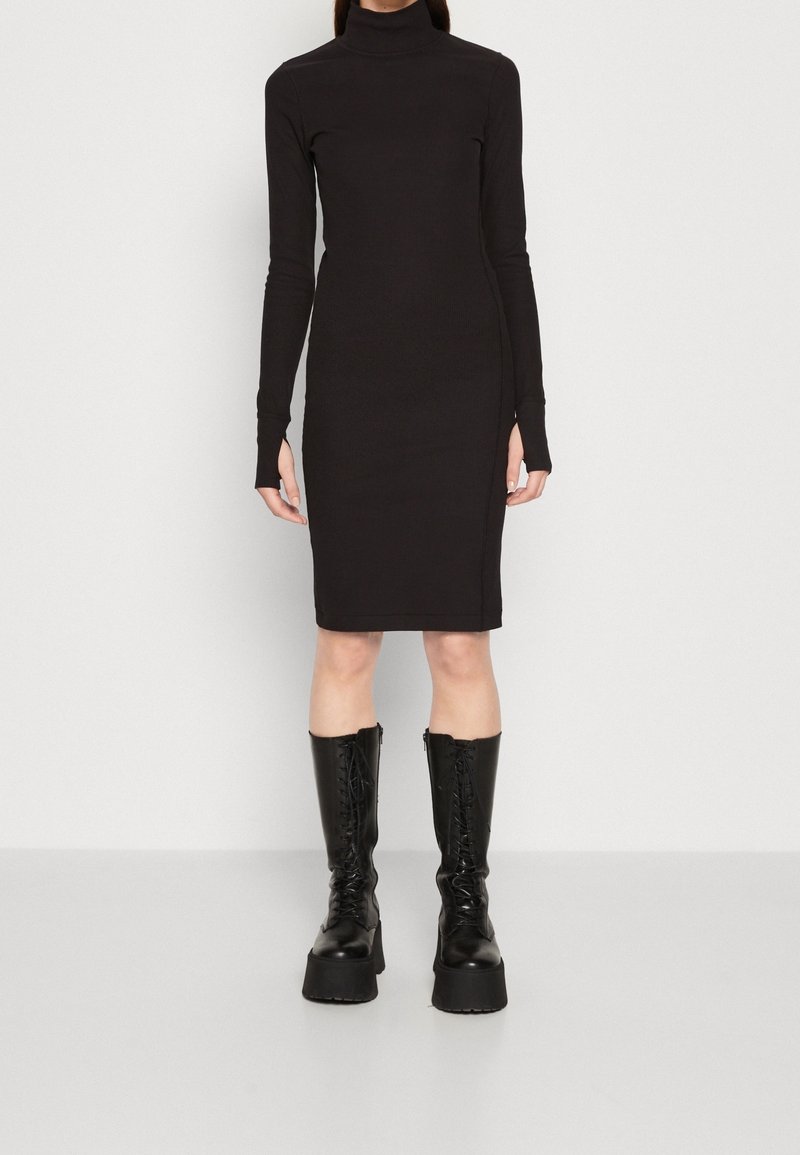Femme portant une robe col roulé noire ajustée à manches longues et des bottes noires à plateforme montant jusqu'aux genoux avec lacets, debout devant un arrière-plan uni.