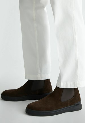 Stivaletti alla caviglia in suede marrone con pannelli elastici laterali e suola in gomma nera, abbinati a pantaloni bianchi a sigaretta con orlo a risvolto.