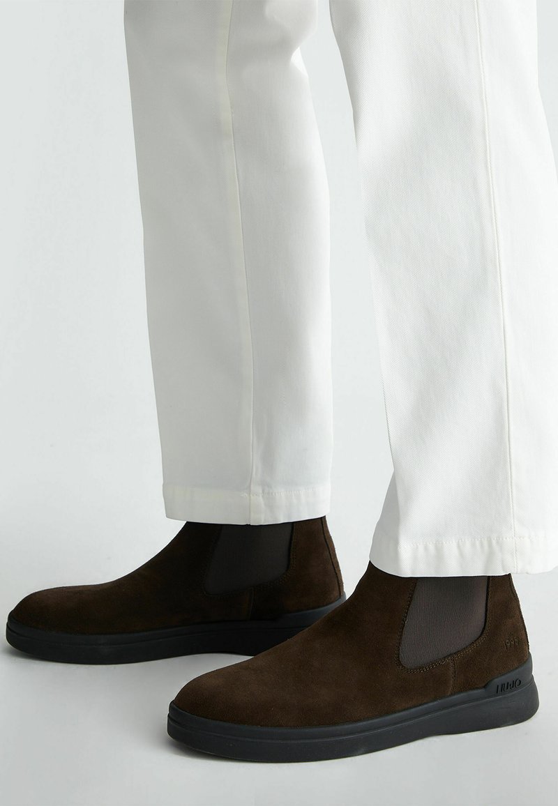 Stivaletti alla caviglia in suede marrone con pannelli elastici laterali e suola in gomma nera, abbinati a pantaloni bianchi a sigaretta con orlo a risvolto.