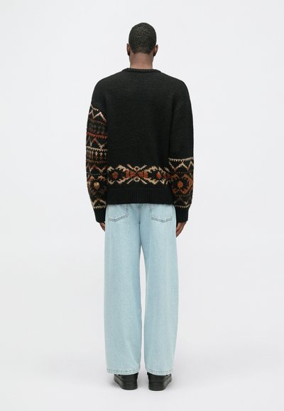 Fekete kötött pulóver, földszínű mintás szegéllyel; laza szabás, kerek nyak, világoskék széles szárú farmernadrággal és fekete cipővel viselve.