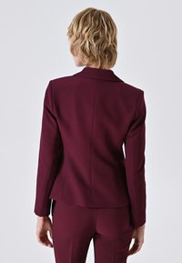 Blazer tailored color bordeaux con rever a punta, silhouette aderente, texture liscia e dettagli con spacco posteriore. Adatto per contesti formali.