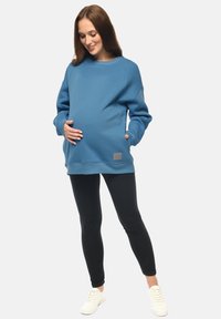 Felpa blu con una vestibilità comoda e maniche lunghe, abbinata a leggings neri e sneakers bianche. Presenta un'etichetta con logo a texture nella parte inferiore.
