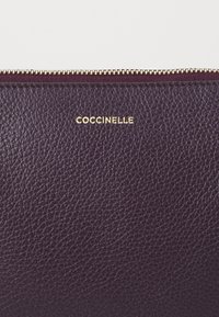 Tumši purpura ādas rokas soma ar teksturētu virsmu, zeltītu rāvējslēdzēju un izspiestu logo "Coccinelle."