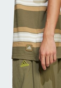 Olīvu zaļš svītrots T-krekls ar brūnām, baltām un bēšām horizontālām svītrām; iekļauj Adidas logo uzlīmi un roku, kas balstās uz atbilstošām šortiem.