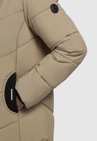 Beige quiltet pufferjakke med lange ærmer, der har et rundt sort logo-patch og elastiske detaljer på lommer og manchetter.