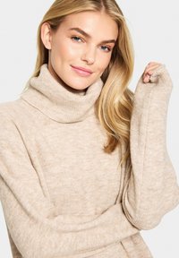 Beige polotröja tillverkad av mjukt, texturerat material med lösa ärmar, som har en ribbad krage och en något oversized passform.