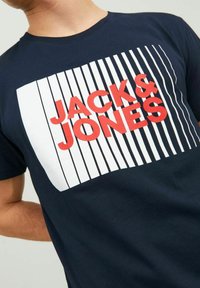 T-shirt albastru marin cu un grafic rectangular alb, având dungi verticale și numele brandului "JACK & JONES" scris cu litere îngroșate roșii.