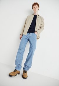 Beige cordfløyels skjorte, mørk marineblå t-skjorte, lyseblå rett ben jeans, og tannfargede sko med svarte såler og snører.