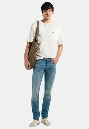 Jeune homme debout, une main dans la poche, portant un t-shirt blanc, un jean bleu, des baskets blanches, une chaîne en argent, et un sac à bandoulière marron.