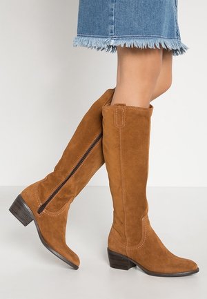 Jambes portant des bottes hautes en daim marron avec fermetures éclair latérales et talons bas, associées à une jupe en denim à ourlet effiloché.