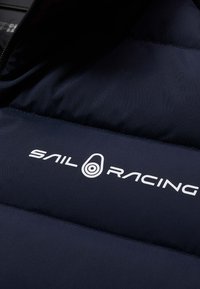 Marine pufferjack met horizontale vullingen, een gladde textuur en een prominent weergegeven witte "SAIL RACING" logo.