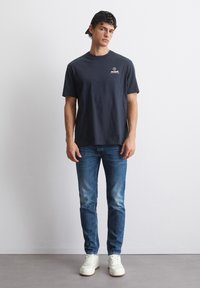 Mørkeblå crew neck T-skjorte med frontlogo, kombinert med lyseblå jeans og hvite sneakers. Modellen har på seg en svart caps.