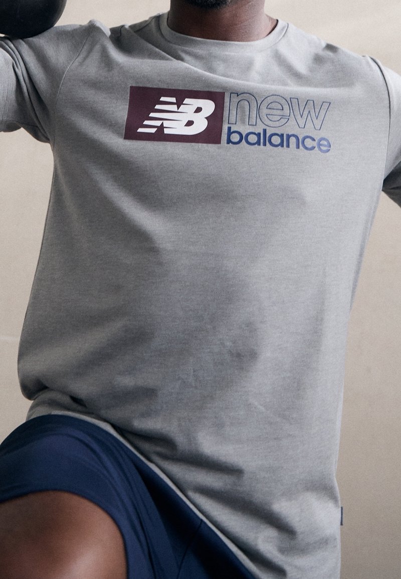 Szara koszulka sportowa z krótkim rękawem z maroonowym i niebieskim logo "New Balance". Miękka tkanina, dopasowany krój, minimalna tekstura.