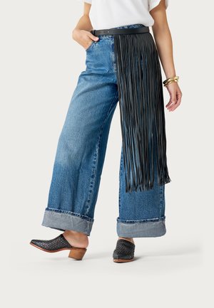 Persona vistiendo jeans anchos con dobladillo, zuecos negros de tacón, una camiseta blanca, cinturón de cuero negro con flecos y pulseras doradas en una muñeca.