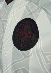 Svart cirkulär lappar med röda "DSU"-bokstäver och "Dansk Boldspil-Union 1889" på grå tyg med abstrakta vita mönster.