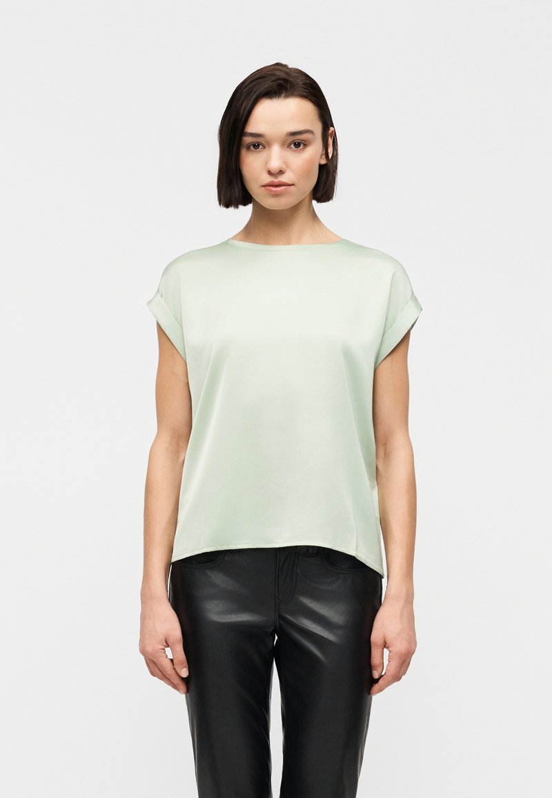 Γυναίκα με κοντά σκούρα μαλλιά, που φοράει ανοιχτό πράσινο μπλουζάκι με κοντά μανίκια και μαύρο δερμάτινο παντελόνι, στέκεται μπροστά σε ένα απλό φωτεινό φόντο.