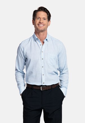 Glimlachende man met donker haar, gekleed in een lichtblauw overhemd met knopen, zwarte broek en een bruine riem, handen in de zakken, staand tegen een witte achtergrond.