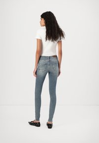 Vaqueros ajustados de denim azul claro con una sutil textura desgastada, cuentan con dos bolsillos traseros y una cinturilla de altura media, combinados con una camiseta blanca.