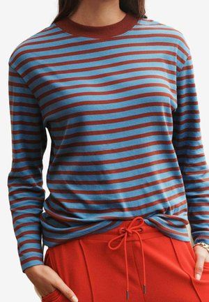 Femme portant une chemise rayée bleu et marron à manches longues, rentrée dans un pantalon rouge à taille à cordon, mains dans les poches.