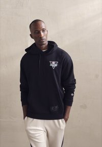 Felpa nera con cappuccio e logo frontale, realizzata in tessuto morbido. Presenta una tasca a marsupio e polsini a costine. Abbinata a pantaloni bianchi.