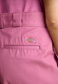 Roze stoffen broek met een achterzak, voorzien van een merklabel met de tekst "Dickies." Het materiaal heeft een gestructureerde afwerking.