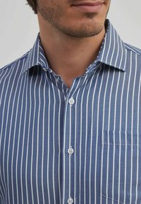 Chemise à rayures bleues et blanches avec col pointu, dotée d'une poche sur le côté gauche et de boutons ronds blancs.