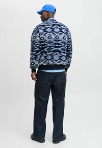 Blauwe en zwarte fleecejack met patroon en hoge kraag, gecombineerd met donkere spijkerbroek en blauwe pet. Gegarandeerd textuurontwerp en ontspannen pasvorm.