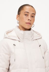 Licht beige gewatteerde jas met een hoge kraag, verstelbare trekkoorden en metalen accenten. Bevat gestikte delen en een capuchon.