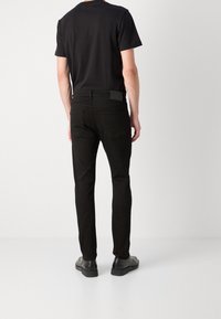 Černé slim fit džíny s kapsami na zadní straně, doplněné černým tričkem. Oblečení zahrnuje černé boty s hladkým povrchem.