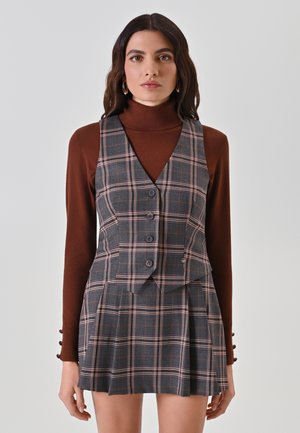 Gilet a quadri grigi con cinque bottoni, indossato sopra un dolcevita ruggine. La minigonna presenta delle pieghe, che si abbinano al motivo a scacchi del gilet.