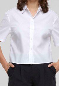 White cropped button-up shirt with a pointed collar, short sleeves, and a smooth texture. Paired with dark, pleated trousers.