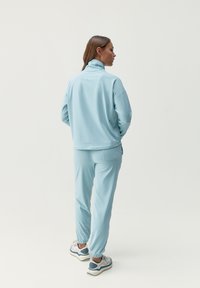 Chándal de tela suave de color azul claro con cuello con cremallera, de corte relajado. Pantalones ajustados con puños elásticos. Zapatillas blancas y grises que complementan el conjunto.