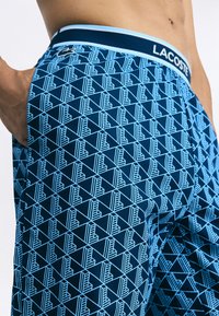 Bermudas de banho masculinas Lacoste com um padrão geométrico em azul claro sobre uma base azul escura, com um cós de contraste e detalhe do logo.