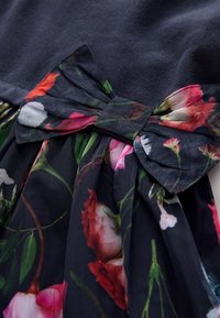 Florales Kleid mit dunklem Oberteil und einer Schleifenapplikation sowie einem fließenden Rock, der mit bunten Blumenmustern auf transparentem Stoff verziert ist.
