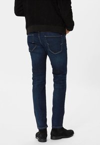 Jeans de mezclilla azul oscuro con un corte ajustado y bolsillos traseros, combinados con una chaqueta negra texturizada y botas negras hasta el tobillo.