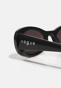 Black sunglasses with glossy finish, wide oval lenses, dark tinted lenses, and engraved "vogue" logo on the side of the sleek arms.