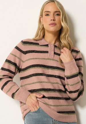 Donna con capelli biondi lunghi che indossa un maglione rosa con collo, righe orizzontali nere e verdi e jeans azzurri.