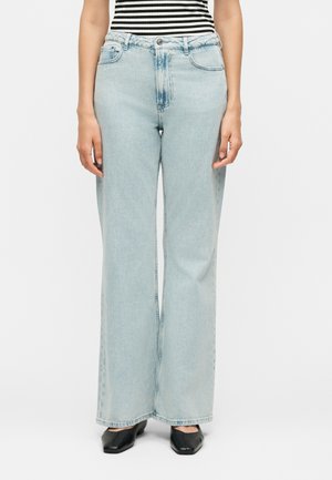 Osoba oblečená v svetlomodrých džínsach s širokými nohavicami, čiernych topánkach s štvorcovou špičkou a čierno-bielom pruhovanom topi, stojaca na bielom pozadí.