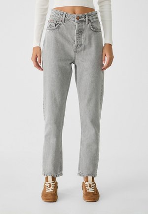 Person iført lysegrå high-waist jeans med lige ben og brune ruskindssneakers med hvide snørebånd, stående mod en ensfarvet baggrund.
