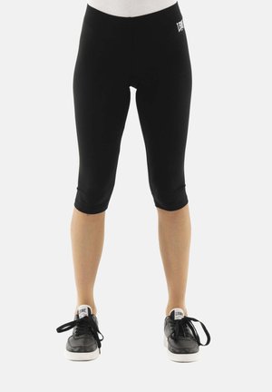 Persona che indossa leggings neri al ginocchio e sneakers nere in piedi su uno sfondo chiaro e uniforme, rivolta in avanti, lacci delle scarpe slacciati.
