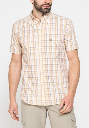 Uomo che indossa una camicia a quadri gialla e beige a maniche corte con pantaloncini cargo beige, in piedi davanti a uno sfondo bianco semplice.