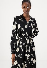 Μαύρο φλοράλ φόρεμα με μακριά μανίκια, κουμπιά μπροστά και ζώνη στη μέση, διακοσμημένο με λευκά αφηρημένα λουλούδια και απαλή υφή υφάσματος.